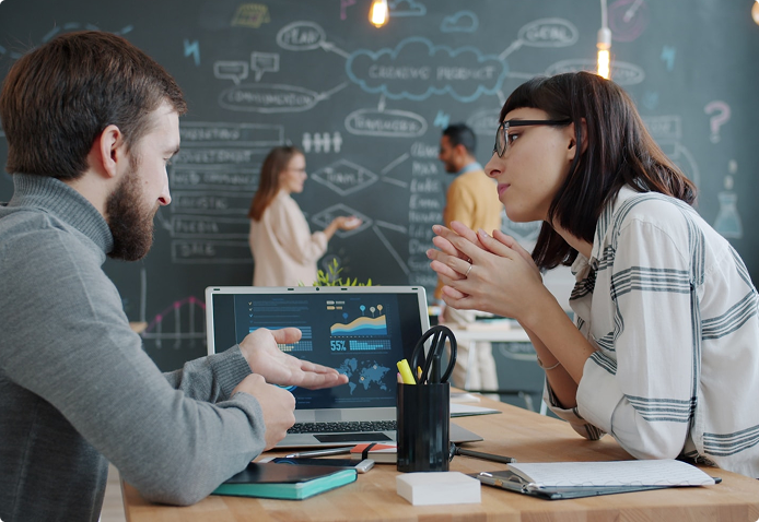 Two people sit at a desk discussing data shown on a laptop screen, with notebooks and office supplies nearby. In the background, two others work in front of a chalkboard covered in charts and diagrams.