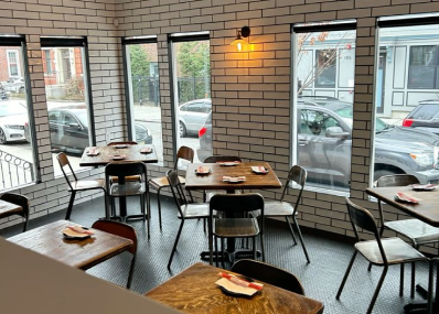 A small restaurant dining area with wooden tables, metal chairs, and white brick walls. Place settings with napkins and utensils are on the tables. Large windows show parked cars and buildings outside.
