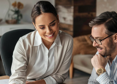 A woman and a man sit together at a desk, looking at documents or a computer screen. The woman is smiling and pointing, while the man, wearing glasses and a watch, looks on with interest.