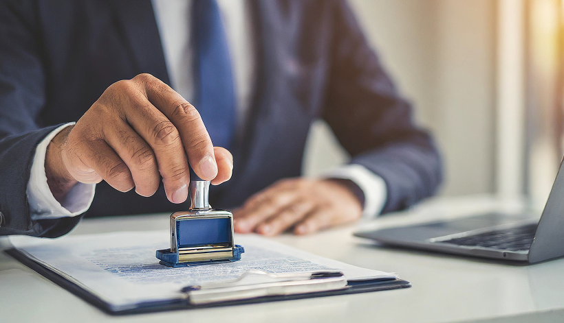 A person in a suit presses a rubber stamp onto a document on a desk, with a laptop and paperwork visible in the background.