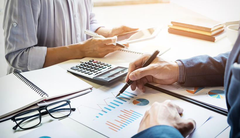 Two people reviewing financial charts and graphs at a desk with a calculator, notepads, and a tablet. One person is writing on a printed chart while the other holds a pen and a tablet.