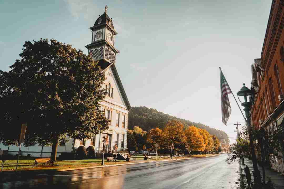 A clock tower stands next to a tree on a quiet street lined with autumn-colored trees and buildings. An American flag hangs from a pole, and morning sunlight reflects off the wet pavement.