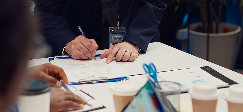A person in business attire is standing at a table, signing a document on a clipboard while another person sits nearby. The table has coffee cups, papers, pens, and office supplies.