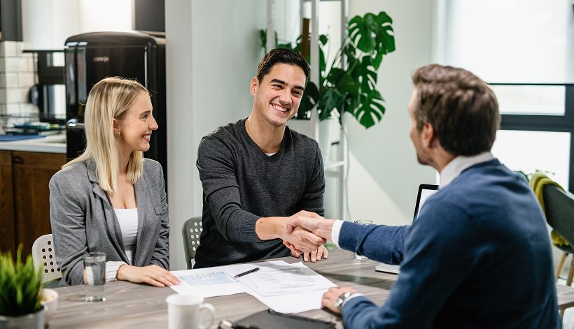 Three people sit at a table in a modern office. Two people, a man and a woman, face a third man. The two men are shaking hands, and paperwork is visible on the table. All appear to be in discussion.