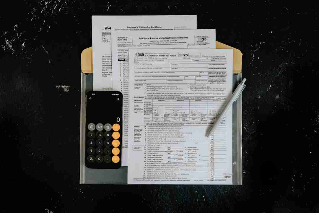 A smartphone with a calculator app open, IRS tax forms, and a pen are arranged on a folder atop a black surface.