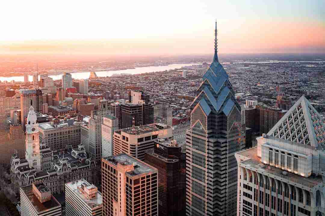 Aerial view of downtown Philadelphia at sunset, showing modern skyscrapers, historic buildings, and the Delaware River in the background under a clear sky.