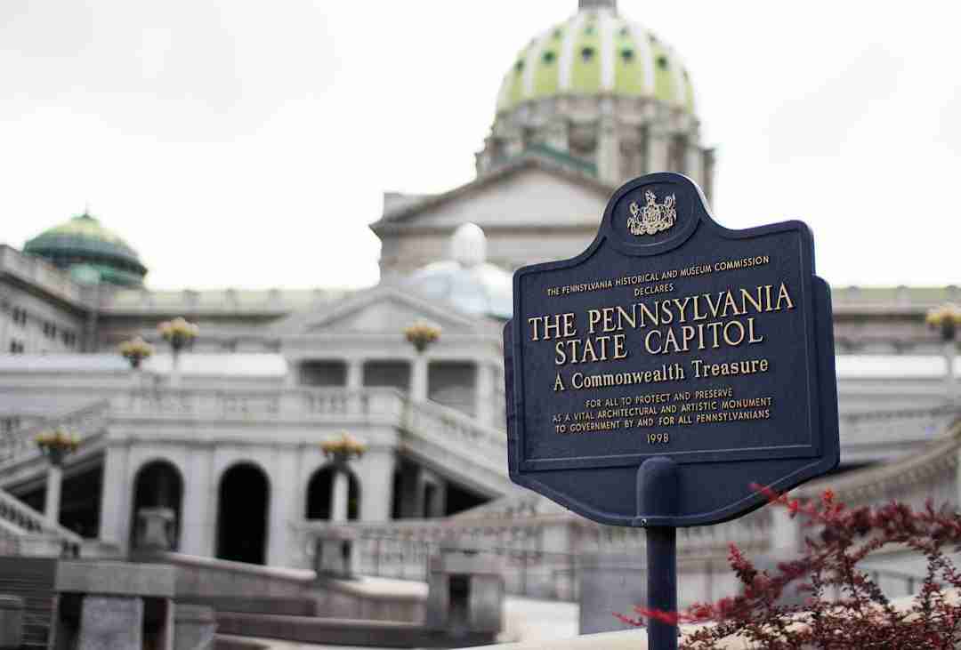 A historical marker sign for The Pennsylvania State Capitol is in focus, with the capitol building's dome and architectural details visible in the background under an overcast sky.