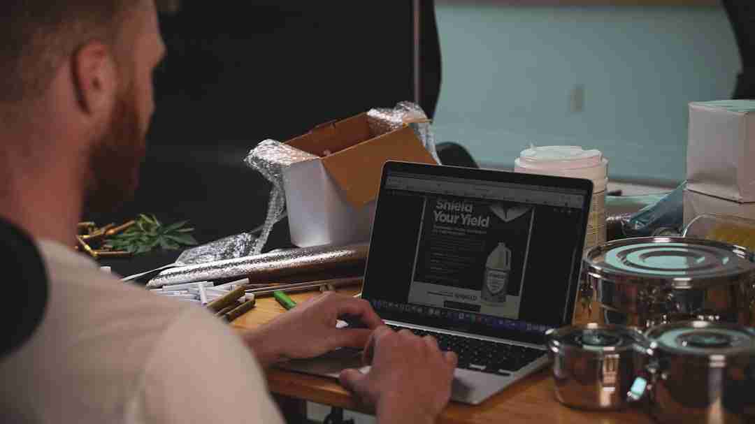 A person sits at a wooden table using a laptop, surrounded by various items including a cardboard box, metal containers, bubble wrap, and a plastic jar. The laptop screen displays a website.
