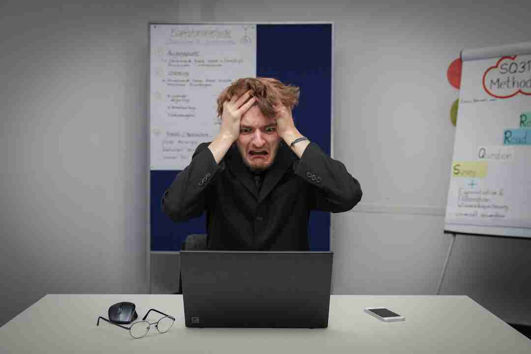 A man in a suit sits at a desk with a laptop, holding his head in frustration. Glasses, a computer mouse, and a smartphone are on the desk. Charts and notes are visible on boards in the background.