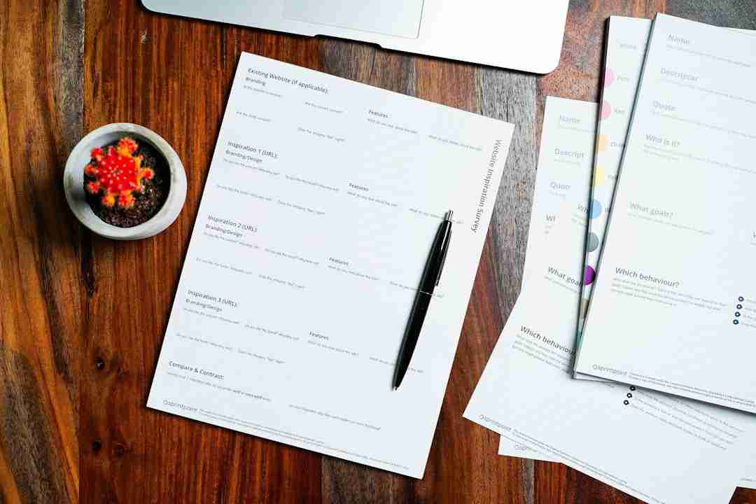 A black pen rests on a printed worksheet atop a wooden table. A small potted cactus and other stacked papers are nearby, with part of a laptop visible in the upper part of the image.