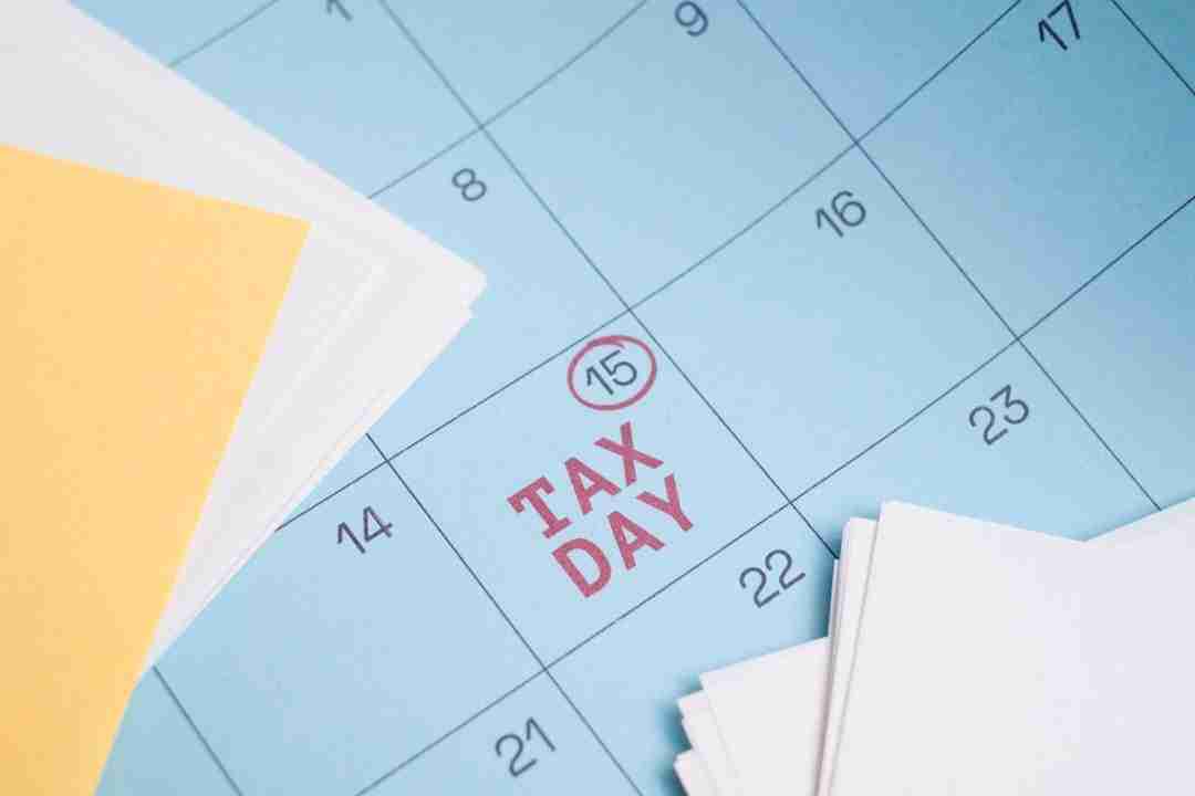 A close-up of a blue calendar with the 15th circled in red and labeled "TAX DAY," surrounded by scattered papers and folders in the foreground.