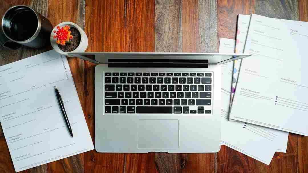 A silver laptop sits open on a wooden desk surrounded by scattered documents, a pen, a cup of coffee, and a small potted cactus.