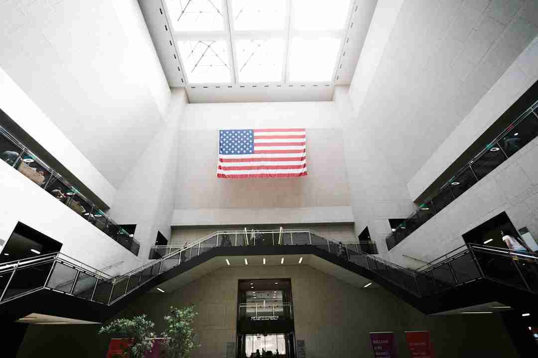 A large American flag hangs on a wall inside a spacious, modern building with high ceilings, skylights, and multiple staircases. There are people visible on the upper walkways and a potted plant below.