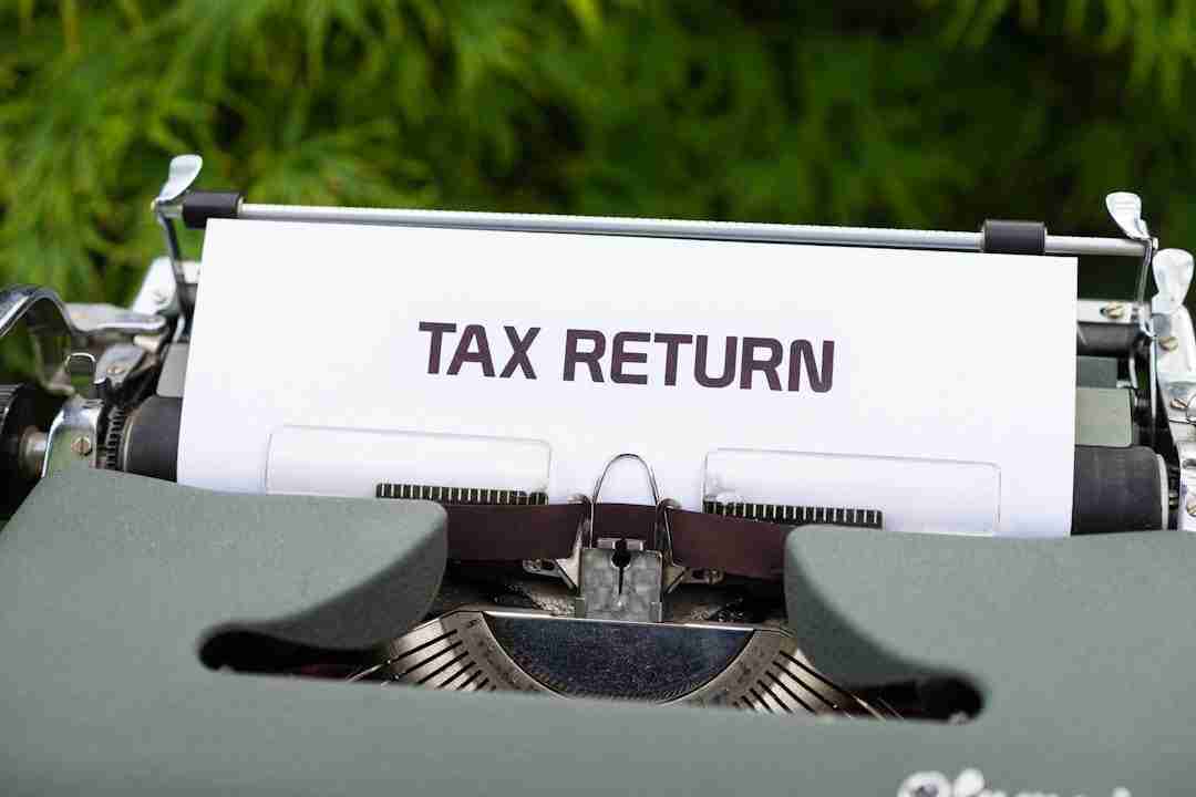 A close-up of a typewriter with a sheet of white paper inserted, displaying the words "TAX RETURN" in bold, uppercase letters. Green foliage is blurred in the background.