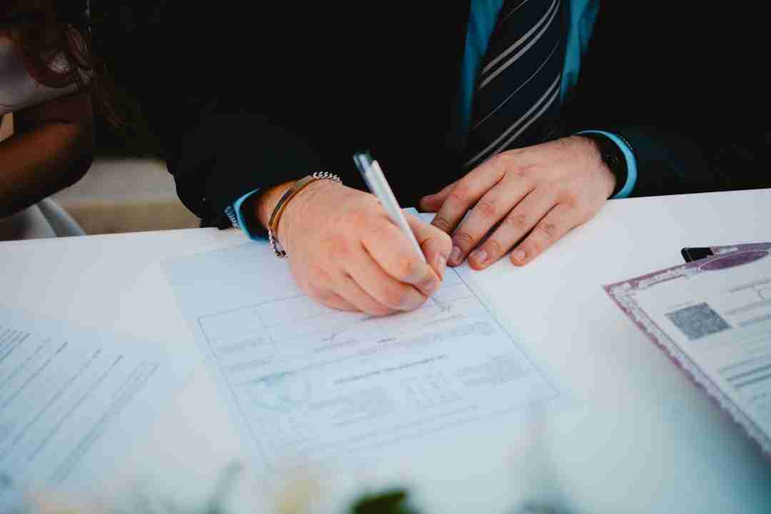 A person wearing a suit and striped tie is signing a document on a white table, with other paperwork visible nearby.