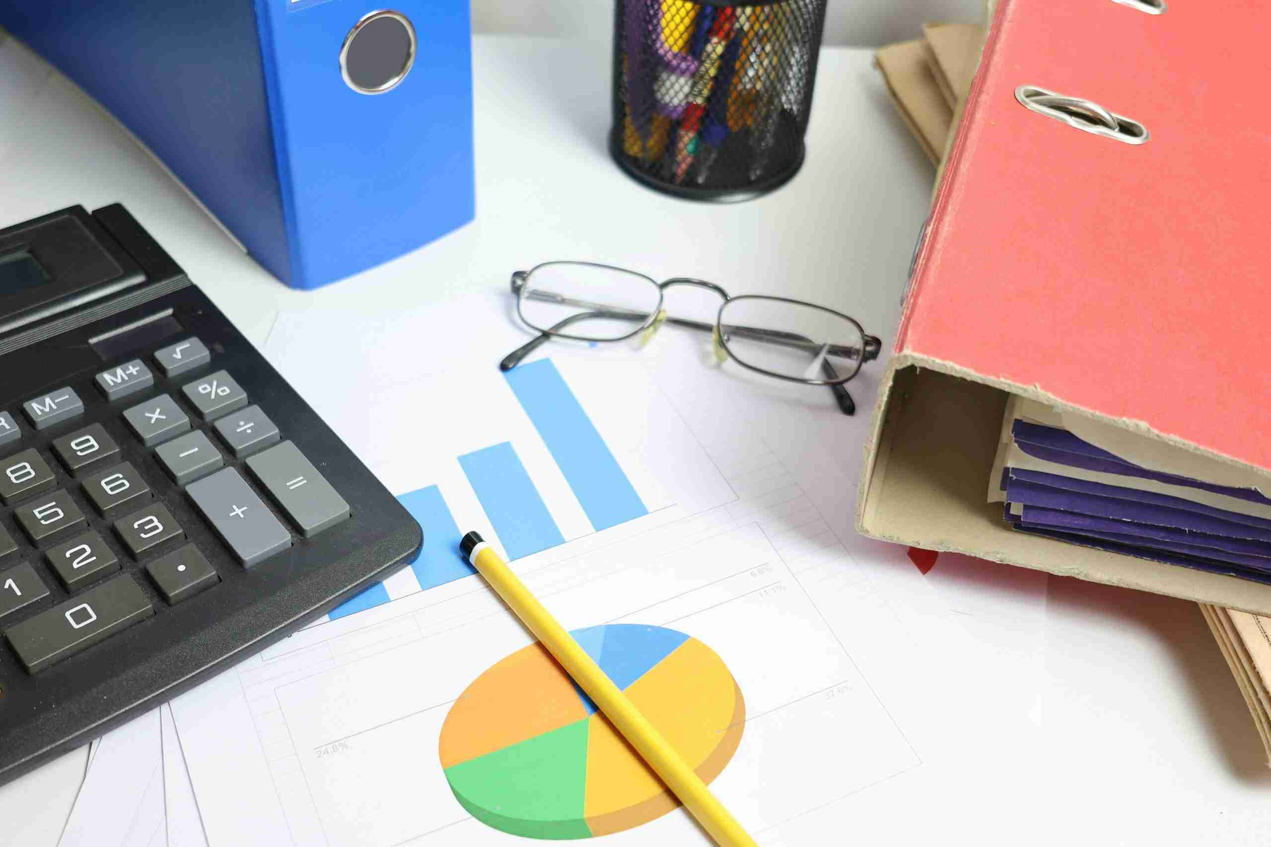 A desk with a calculator, pencil, eyeglasses, bar and pie chart documents, a blue binder, a red file folder filled with papers, and a pen holder with pens and scissors.