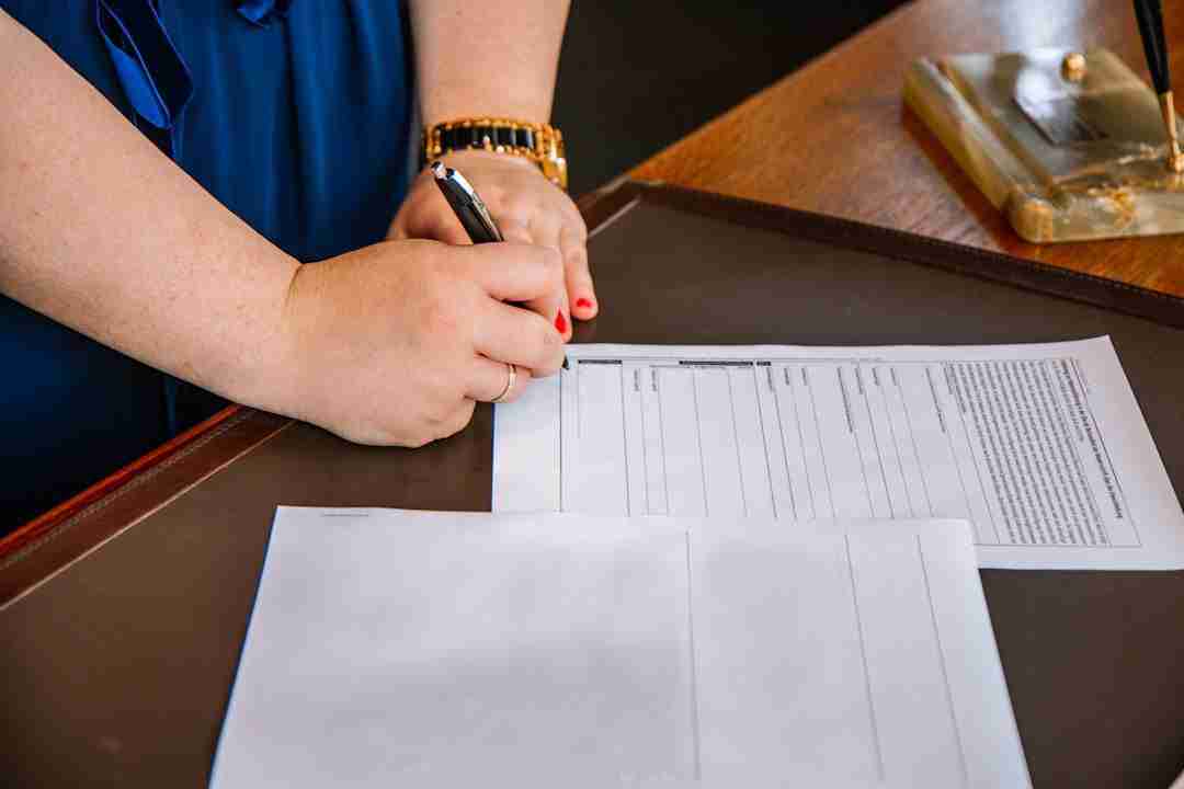 A person wearing a blue outfit and a gold watch is signing a document on a desk. Another sheet of paper and a pen holder are also visible on the desk.