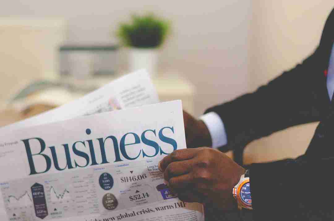 A person in a suit holds a newspaper with the headline "Business." A chart, numbers, and articles are visible on the newspaper. A blurred green plant sits on the desk in the background.
