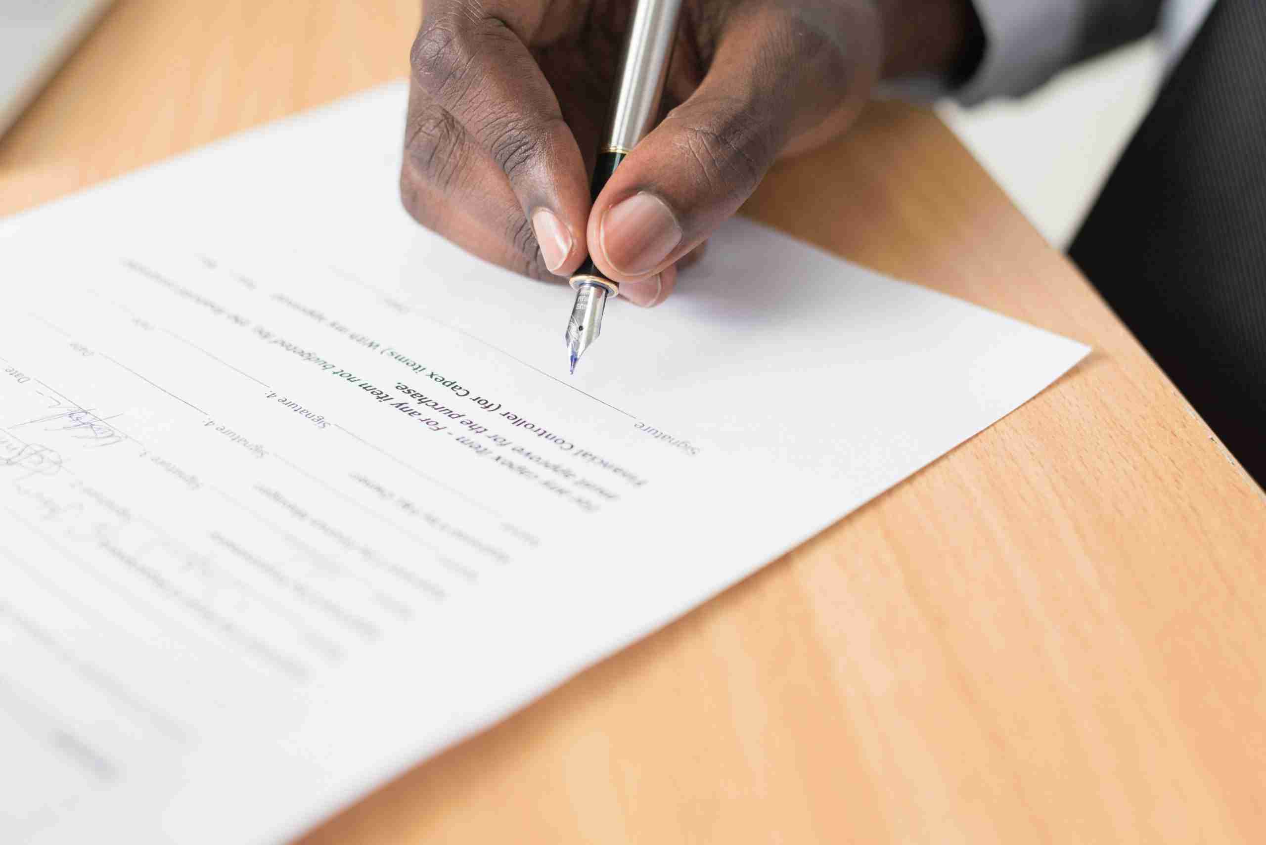 A person’s hand holding a pen is signing a document on a wooden desk. The document contains printed text and several blank lines for writing.