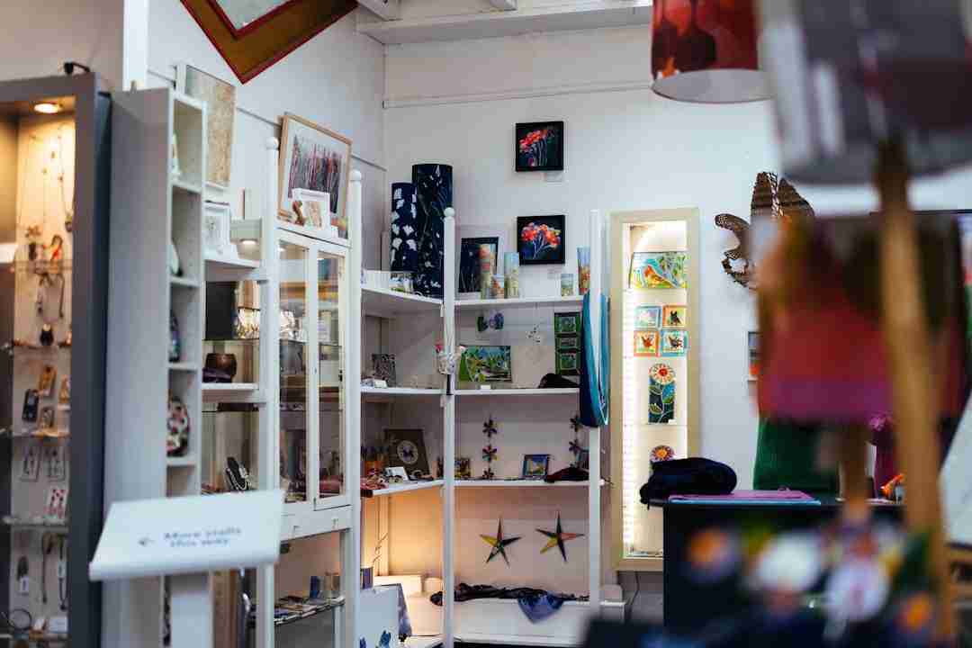 A small art gallery or gift shop interior displays pottery, framed artwork, jewelry, and small crafts on white shelves and cabinets. The space is well-lit with a mix of colorful items and decorations.