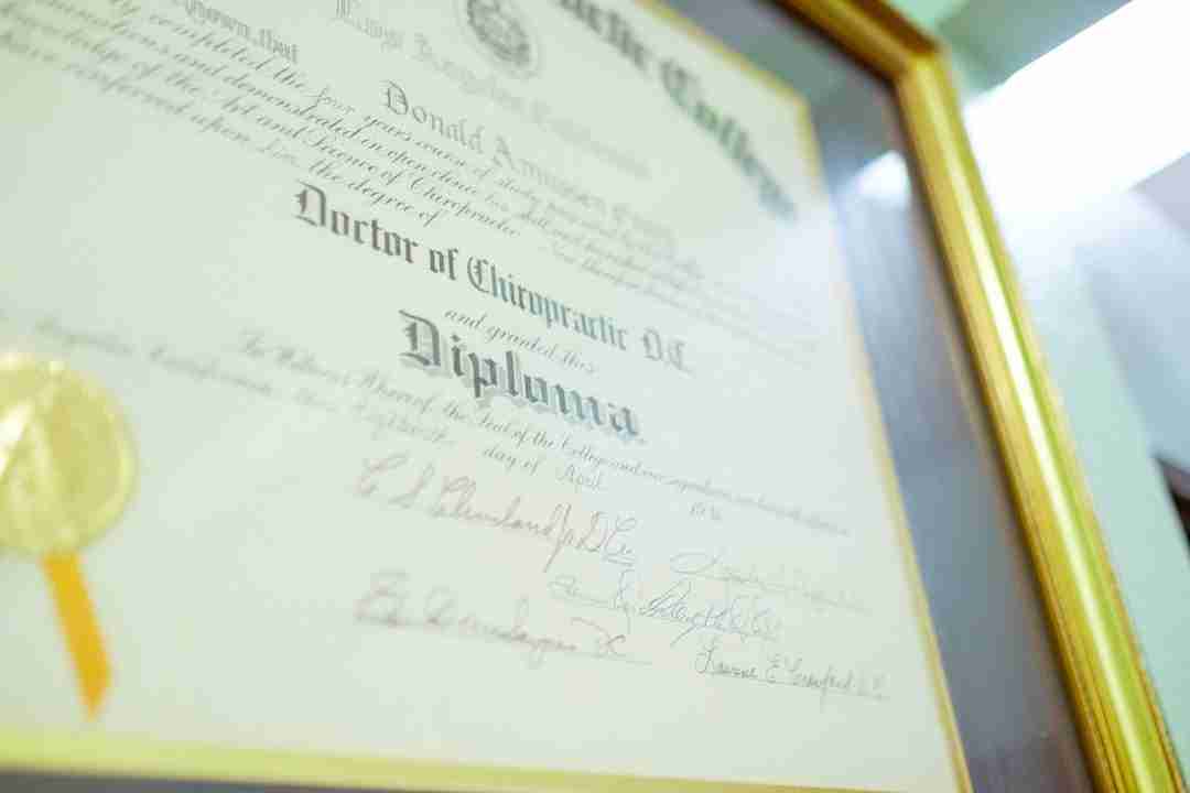A framed Doctor of Chiropractic diploma with signatures and a gold seal, displayed against a white wall. The text on the diploma is partially readable and a gold frame surrounds the document.