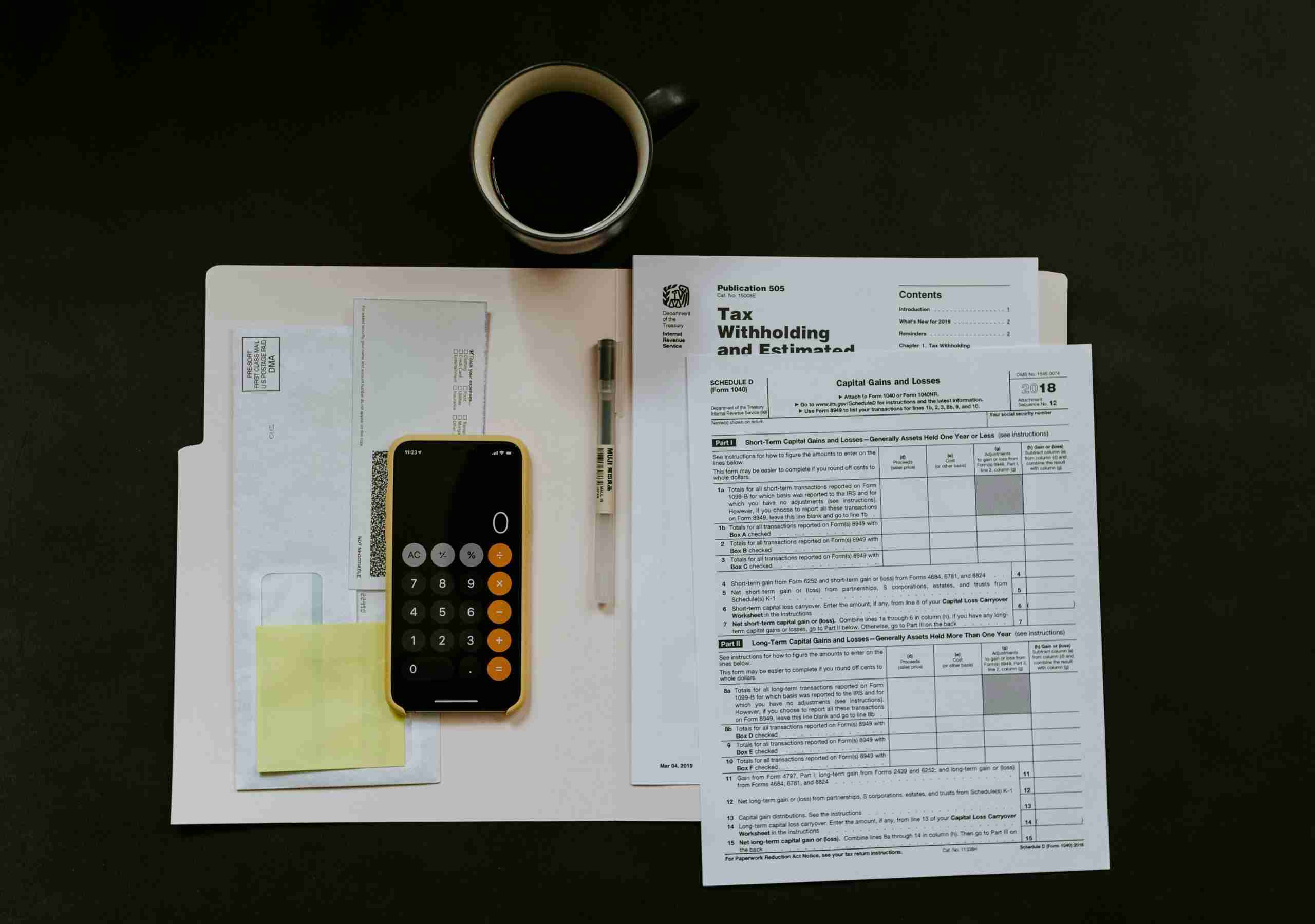 A folder with tax forms, a pen, a smartphone with a calculator app open, an envelope, sticky notes, and a cup of black coffee are arranged on a dark surface.