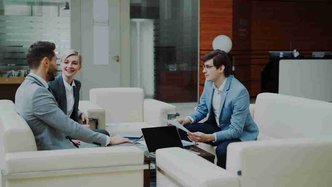 Three people in business attire sit on white sofas around a coffee table with a laptop and documents, engaged in a discussion in a modern office setting.