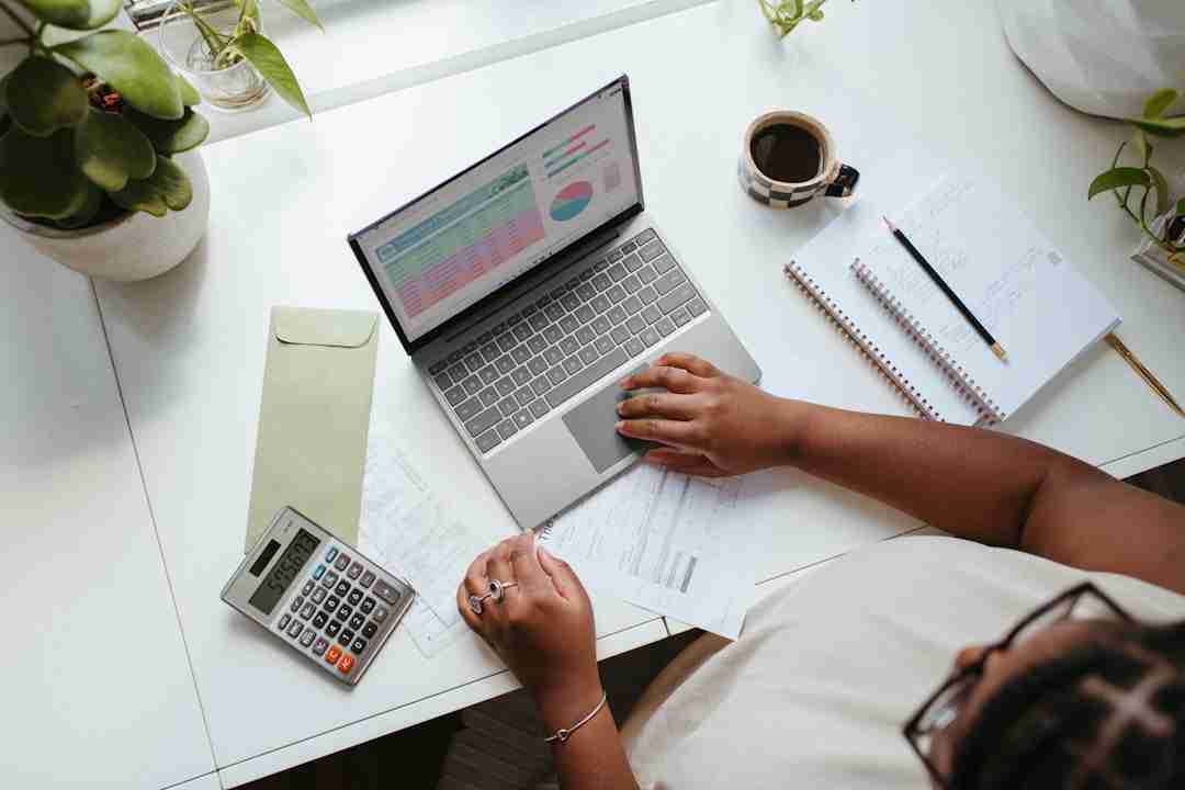 A person working at a desk with a laptop displaying charts, calculator, coffee cup, notepad, papers, and plants nearby. The person is using a mouse and holding a document.