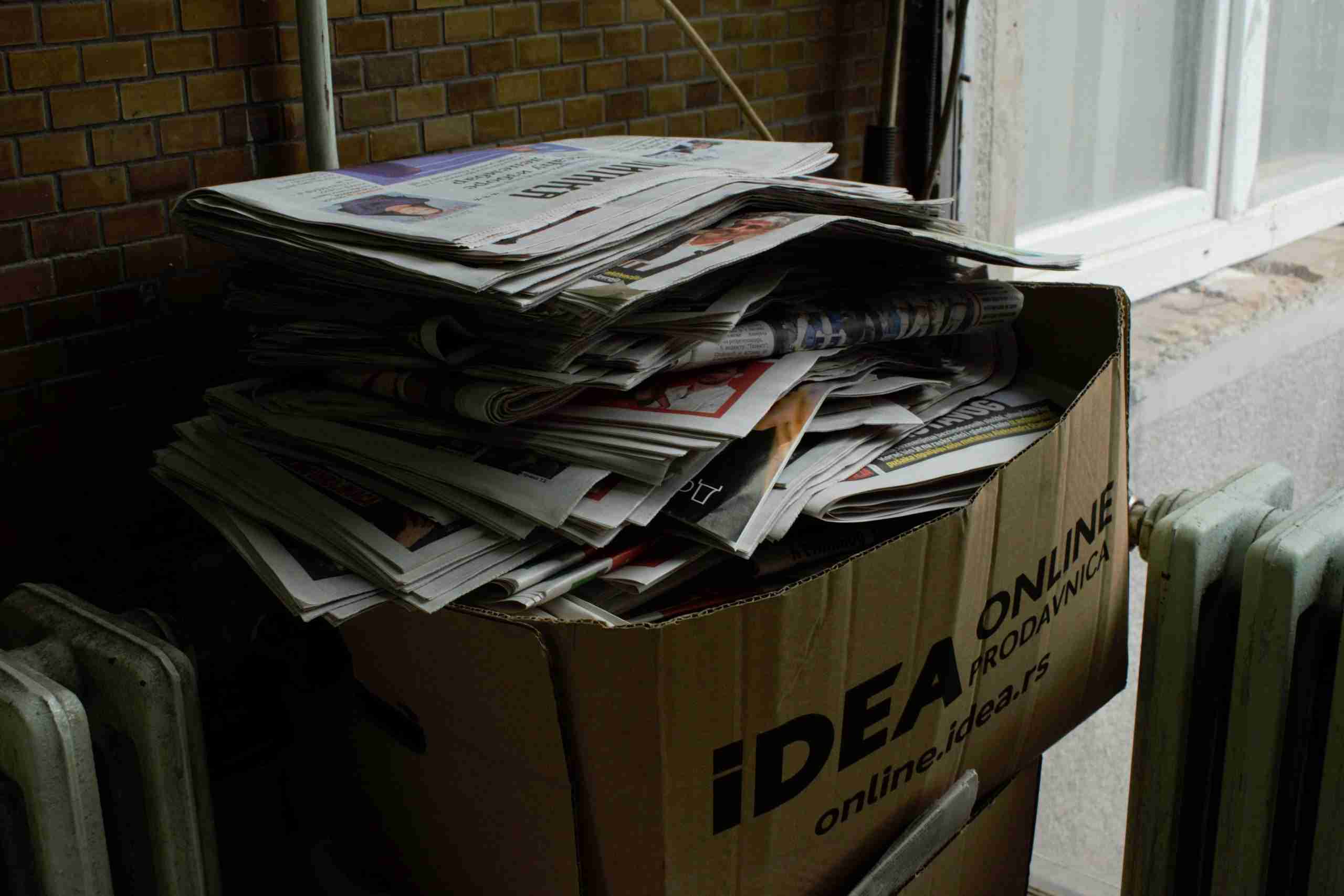 A cardboard box labeled “IDEA ONLINE” is filled with a large, disorganized stack of newspapers and magazines. The box is placed indoors beside a radiator and a brick wall.