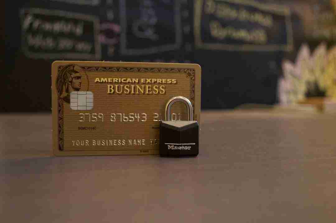 A gold American Express Business credit card stands upright on a table with a small black padlock placed in front of it. A blurry chalkboard is visible in the background.