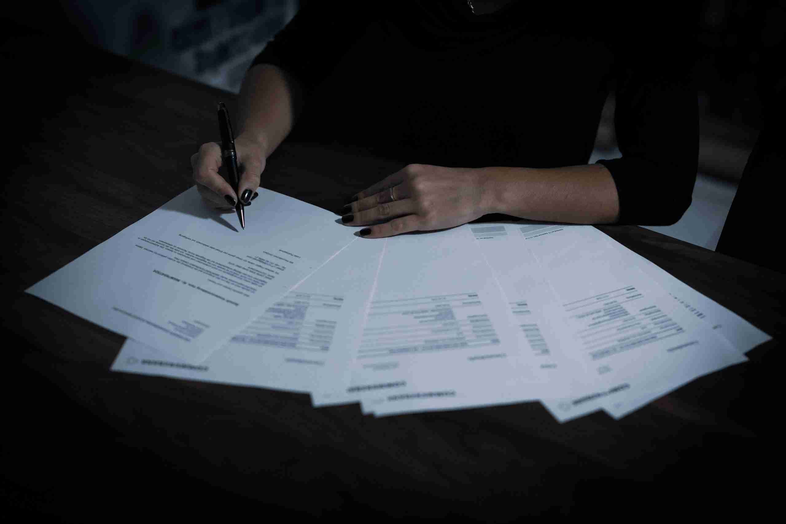 A person with dark nail polish is sitting at a desk, holding a pen and filling out or signing documents spread out on the wooden surface. The image is dimly lit.