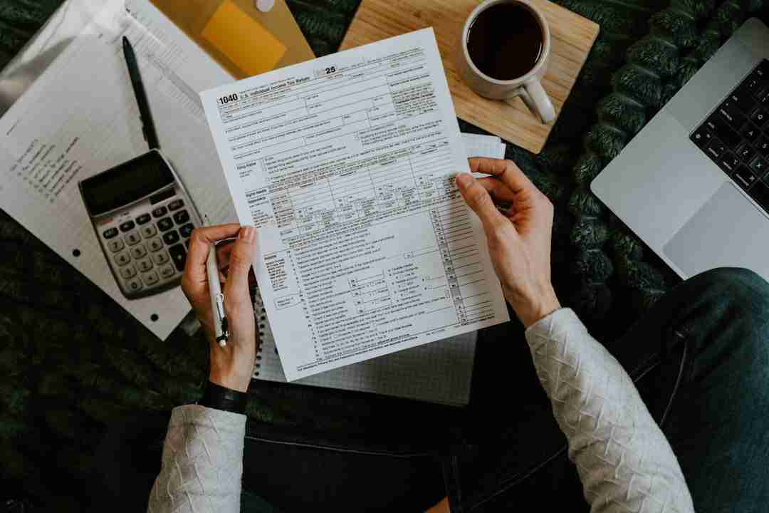 A person holds a completed IRS 1040 tax form with a pen in one hand. Nearby are a calculator, notebook, cup of coffee on a wooden tray, some papers, and part of an open laptop.