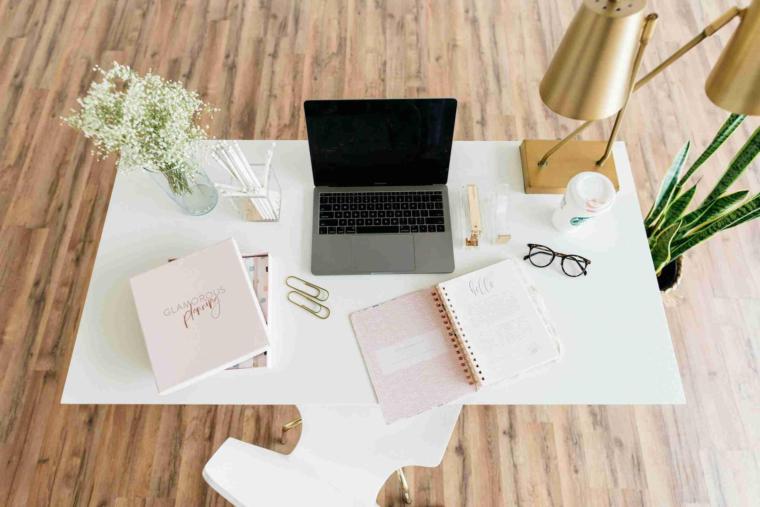 A tidy desk with a laptop, planner, glasses, lamp, coffee cup, plant, gold paperclips, a book, and a vase of flowers on a white surface, set on a wooden floor.
