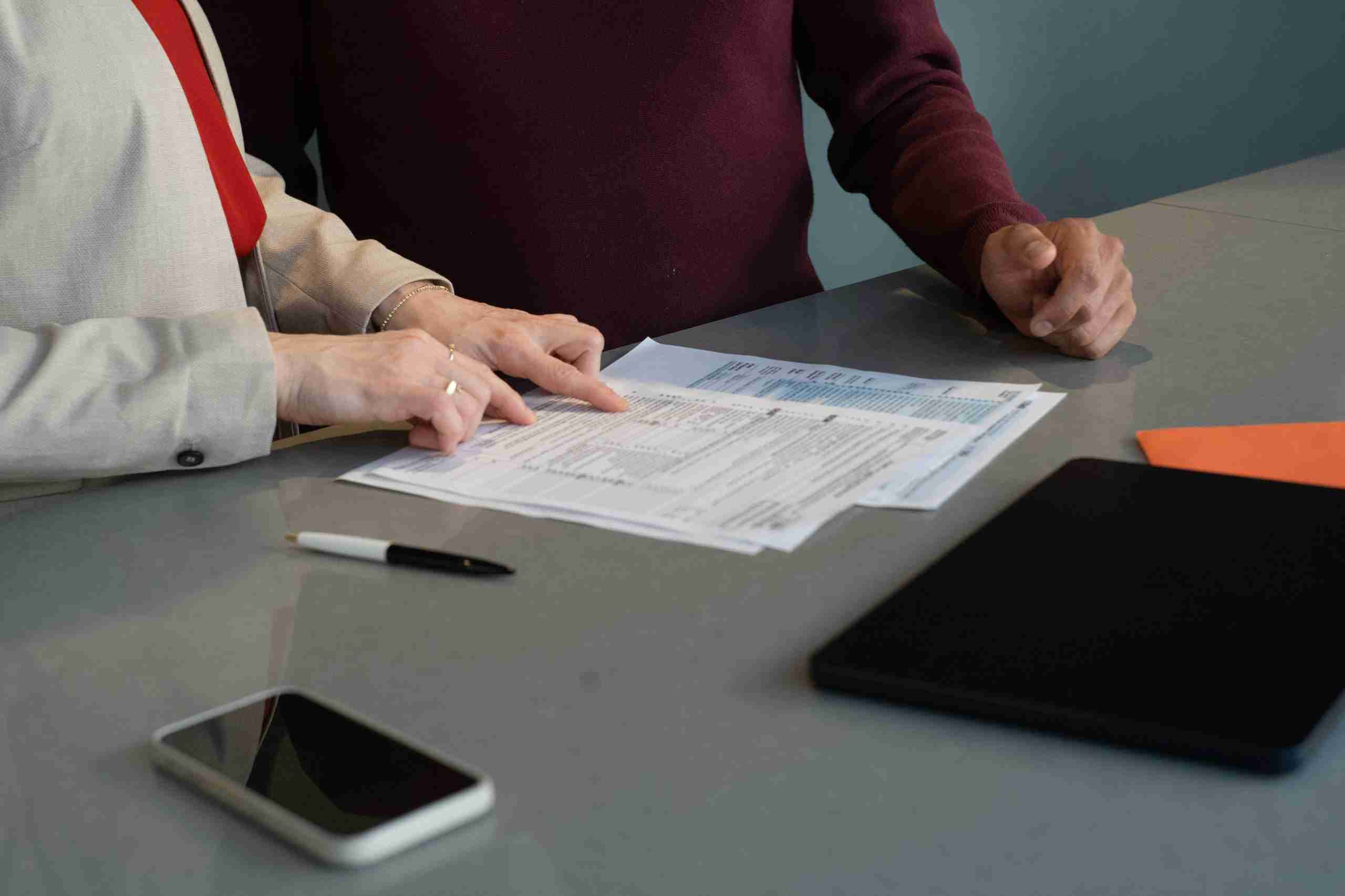 Two people stand at a table reviewing documents, with one person pointing at the papers. A pen, a smartphone, a closed laptop, and an orange folder are also on the table.