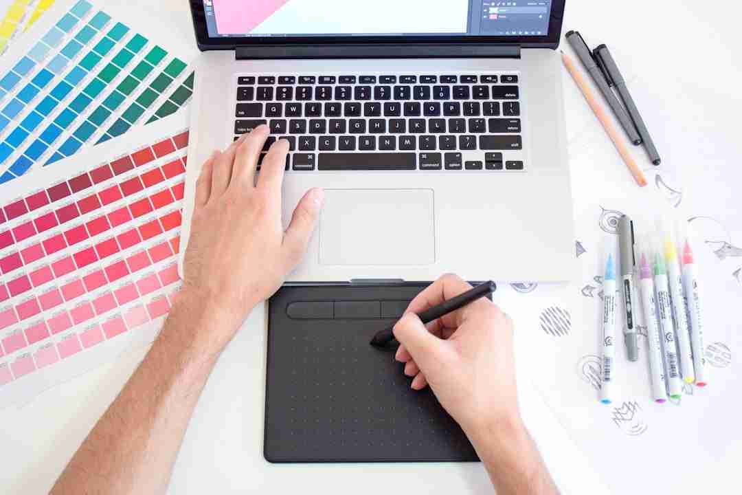 A person uses a graphic tablet and stylus in front of a laptop. Color swatches, markers, pens, and design sketches are on the white desk, suggesting a digital art or graphic design workspace.
