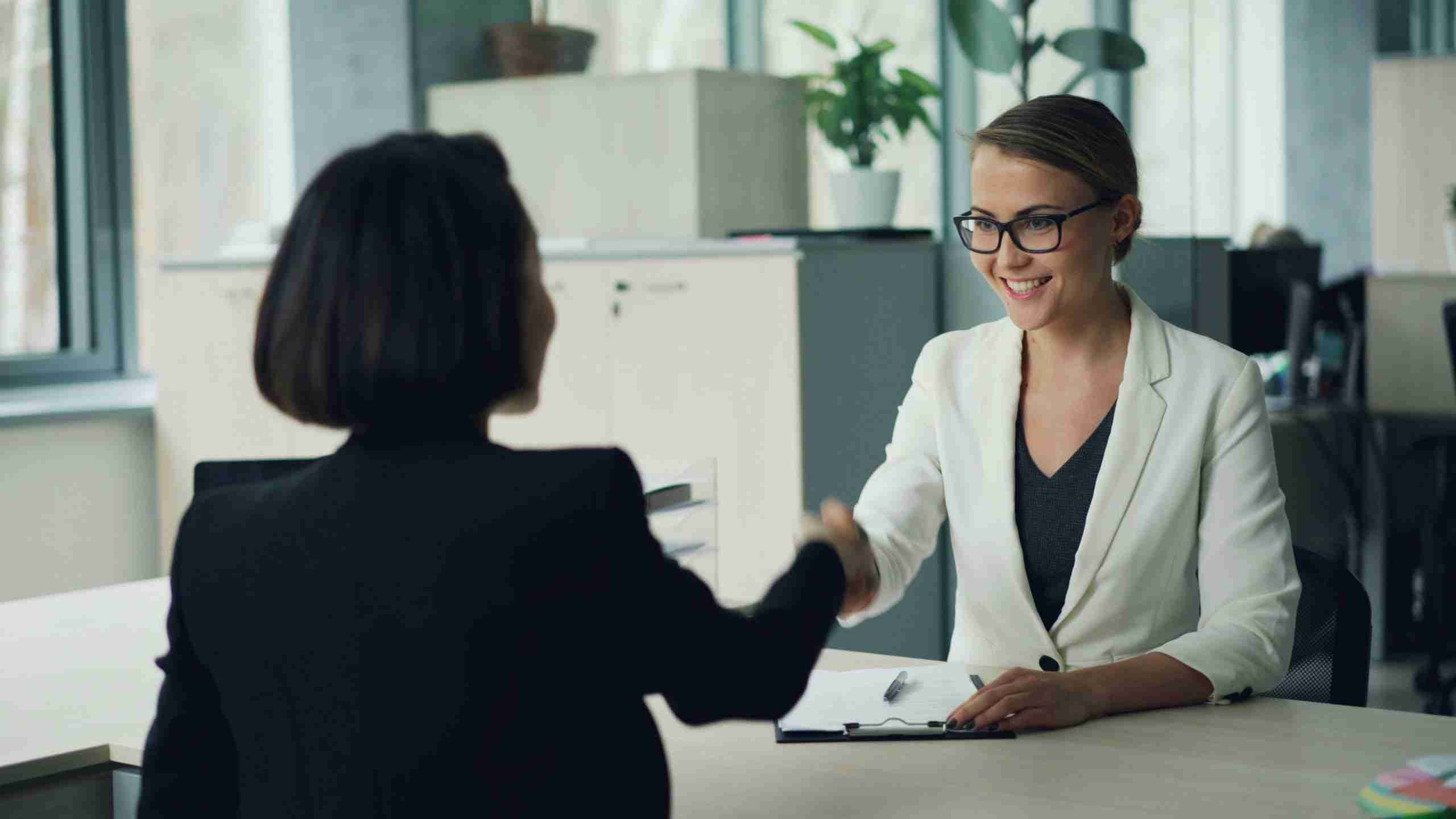 Two women in business attire shake hands across a desk in an office setting, with one woman smiling. There are documents and a pen on the desk, and plants in the background.