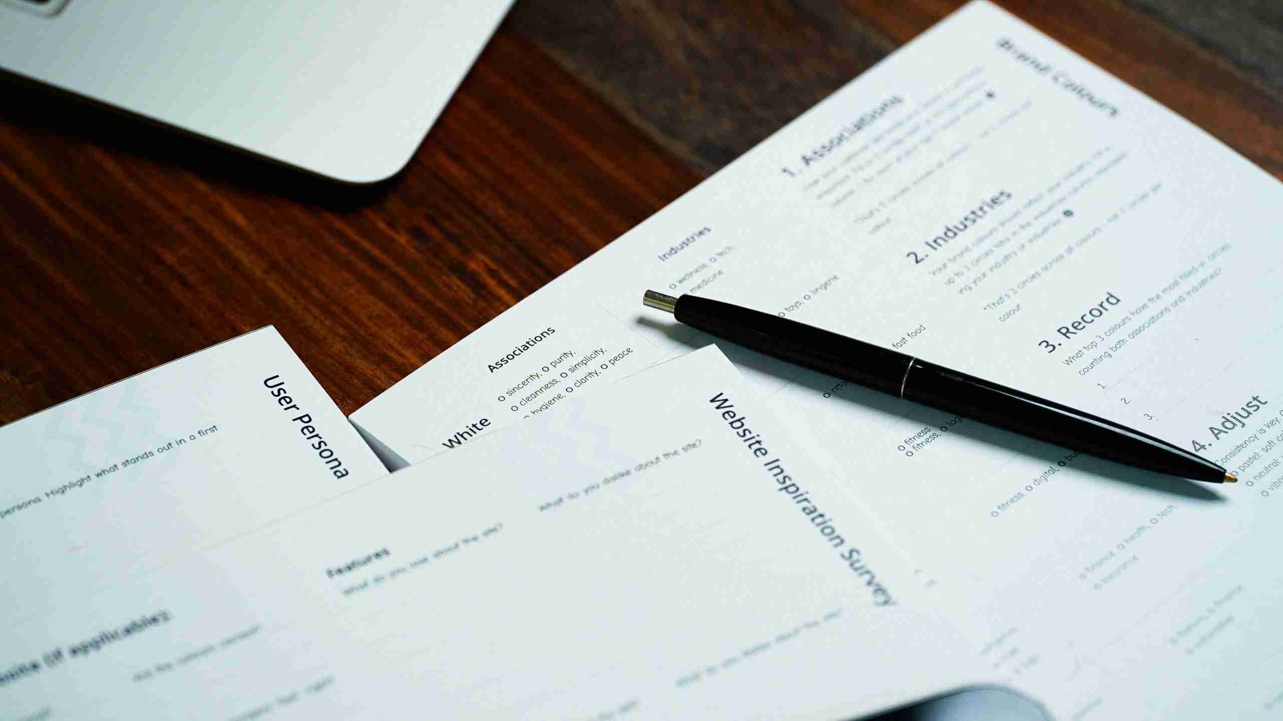A black pen rests on top of printed documents labeled “Website Inspiration Survey” and “User Persona” on a wooden desk, partially visible laptop in the top corner.